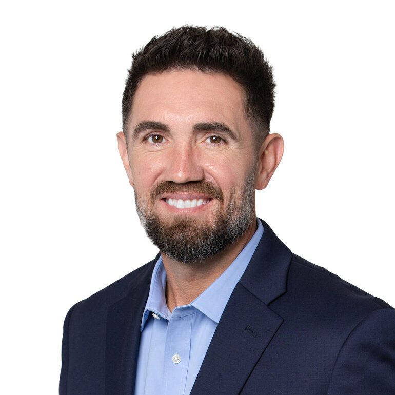 Man with short dark hair and beard, wearing a blue suit jacket and open-collared light blue shirt, smiling.