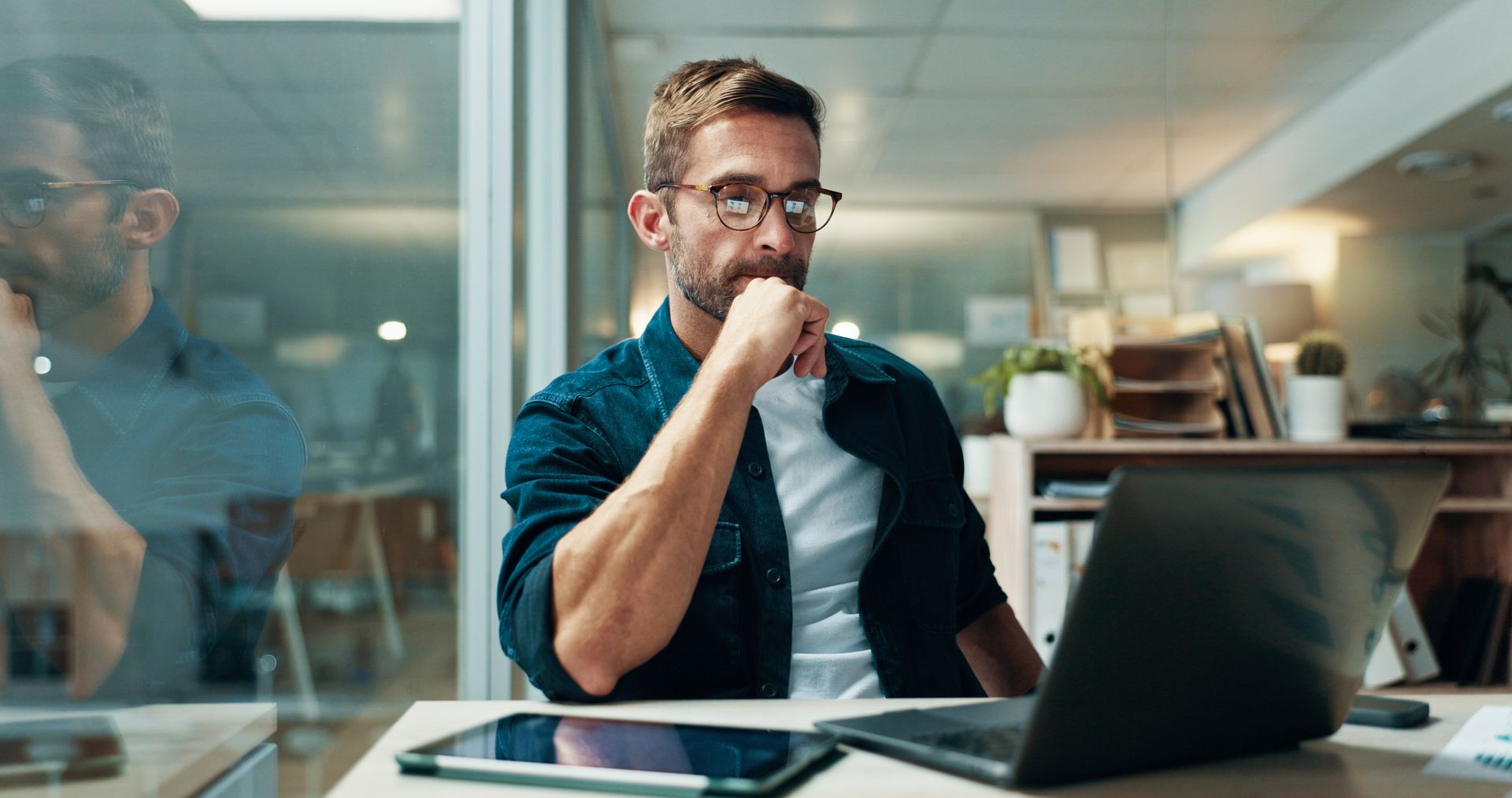 Man in glasses sits at a desk, looking thoughtful at a laptop in a modern office.