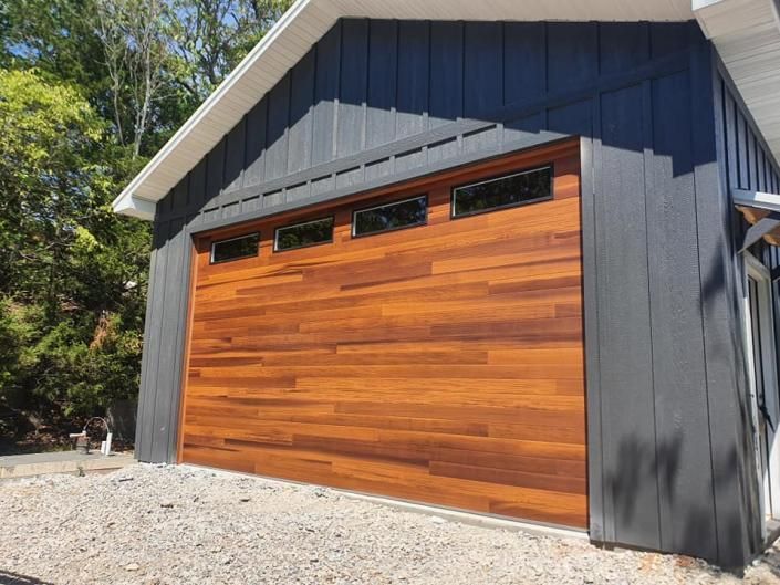 Wood-paneled garage door on a dark gray outbuilding with gravel driveway and trees nearby