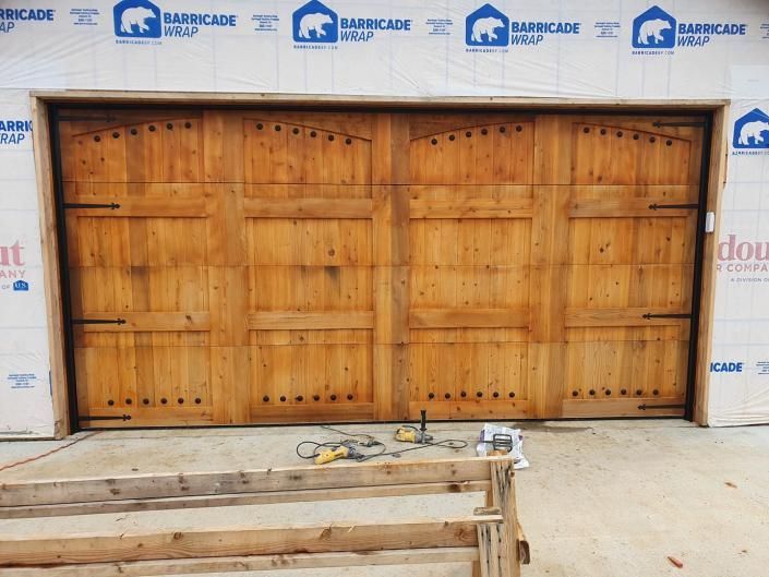 Wooden double garage door framed by construction wrap, with lumber and tools on the ground