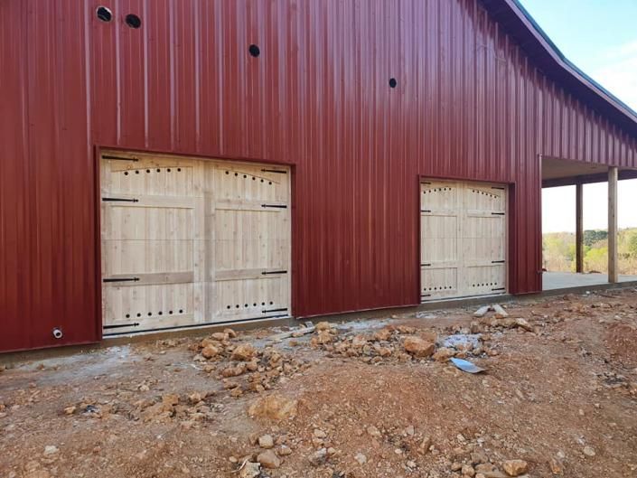 Red barn with two white garage doors on a dirt lot, viewed from the corner.