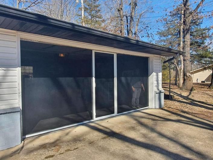Screened porch attached to a gray house, with sliding glass doors and wooded yard beyond.