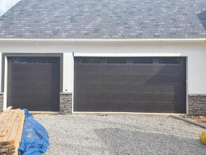 Three-car garage with dark doors, gravel driveway, and stacked lumber covered by a blue tarp on the left.