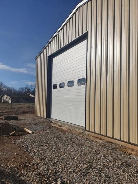 Large beige metal garage with a white roll-up door and gravel driveway under a blue sky