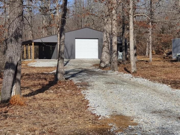 Small gray shed with white garage door at the end of a gravel driveway in a wooded yard