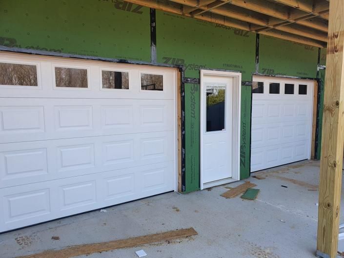 White garage doors and a side entry door on a green building under construction, with unfinished porch posts.