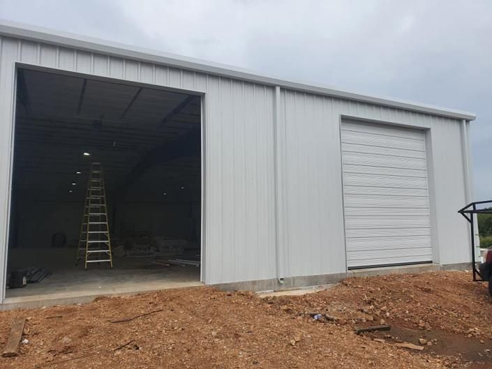 Large gray metal warehouse with an open bay door, closed roll-up door, and a ladder inside on a dirt lot.