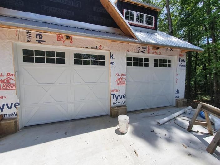 Two white garage doors on a house under construction, with exposed roof framing and construction materials nearby.