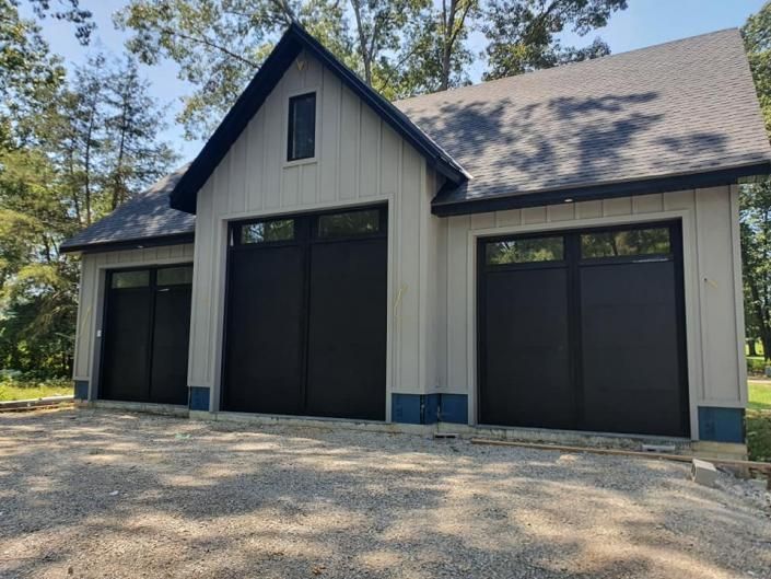 Detached gray garage with three black doors and a gravel driveway, surrounded by trees