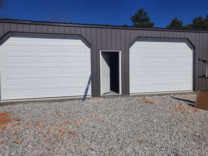 Gray building with two white garage doors and a small open door in the center, on a gravel lot under blue sky