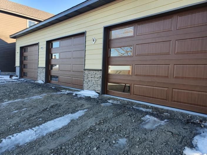 Three brown garage doors on a driveway beside a beige house with snow patches