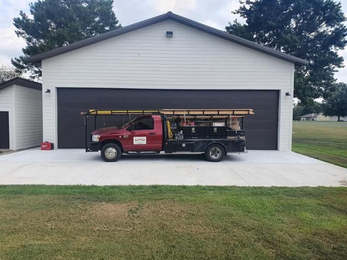 Pickup truck with ladder parked in front of a gray two-car garage with a grassy yard.