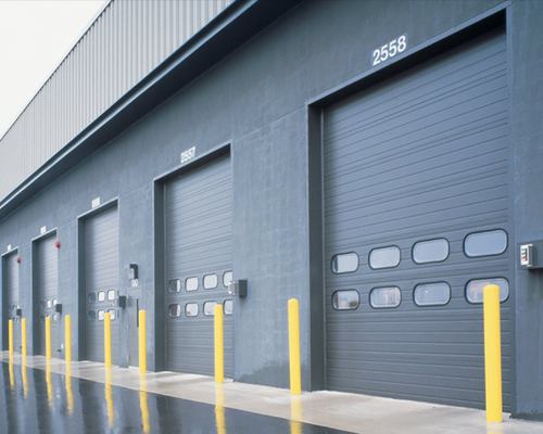 Row of gray warehouse loading bay doors with yellow safety bollards along the dock