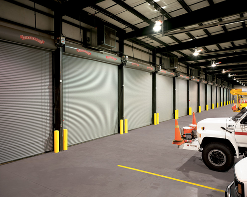 Industrial warehouse with closed roll-up doors, yellow bollards, and a white utility truck inside.