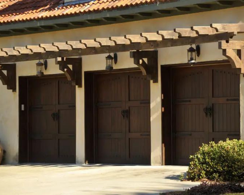 Three brown garage doors under a stucco roof, with lanterns and a shrub at right.