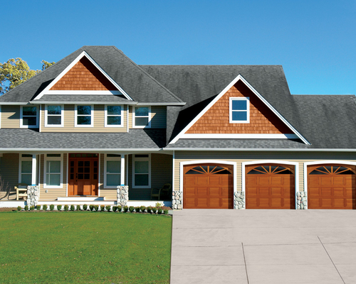 Large suburban house with gray siding, brown garage doors, and a manicured green lawn.