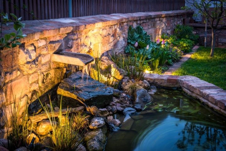 Stone garden waterfall lit at dusk beside a pond and lush landscaping