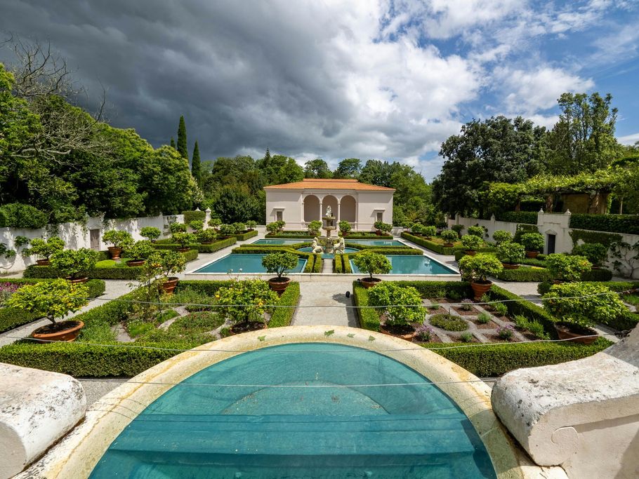 Formal garden with turquoise reflecting pools, manicured hedges, and a small villa under stormy clouds