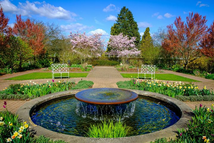 Formal garden with circular fountain, white benches, flower beds, and autumn trees under a blue sky