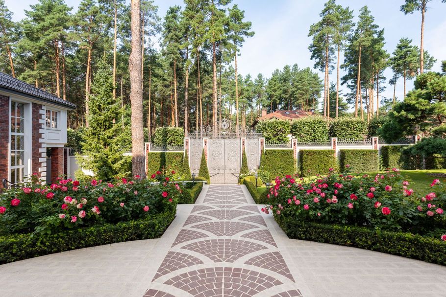 Landscaped garden walkway with pink roses, brick path, and tall pine trees
