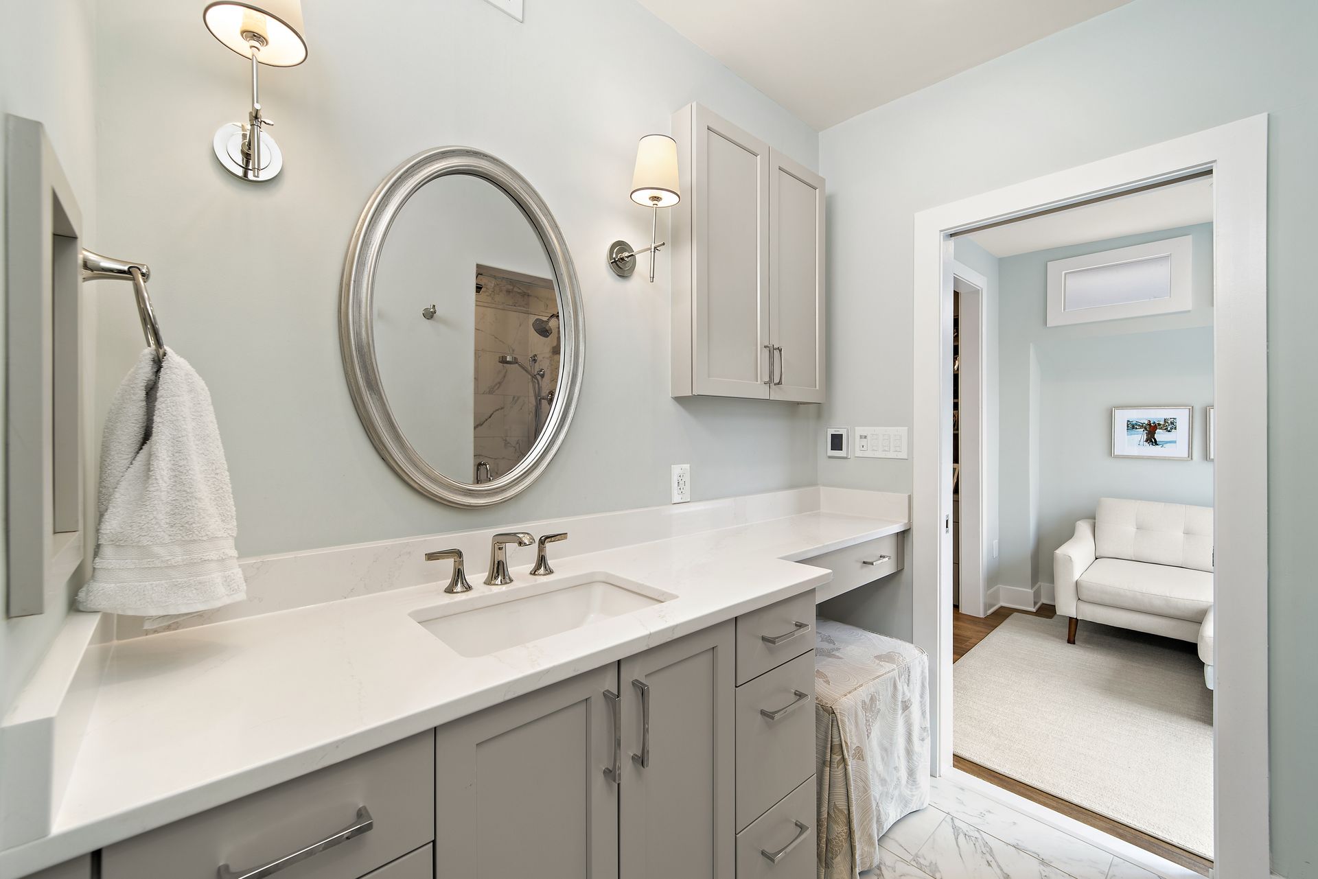Bathroom with gray vanity, marble top, round mirror, sconce lighting, and view into sitting room with white chair