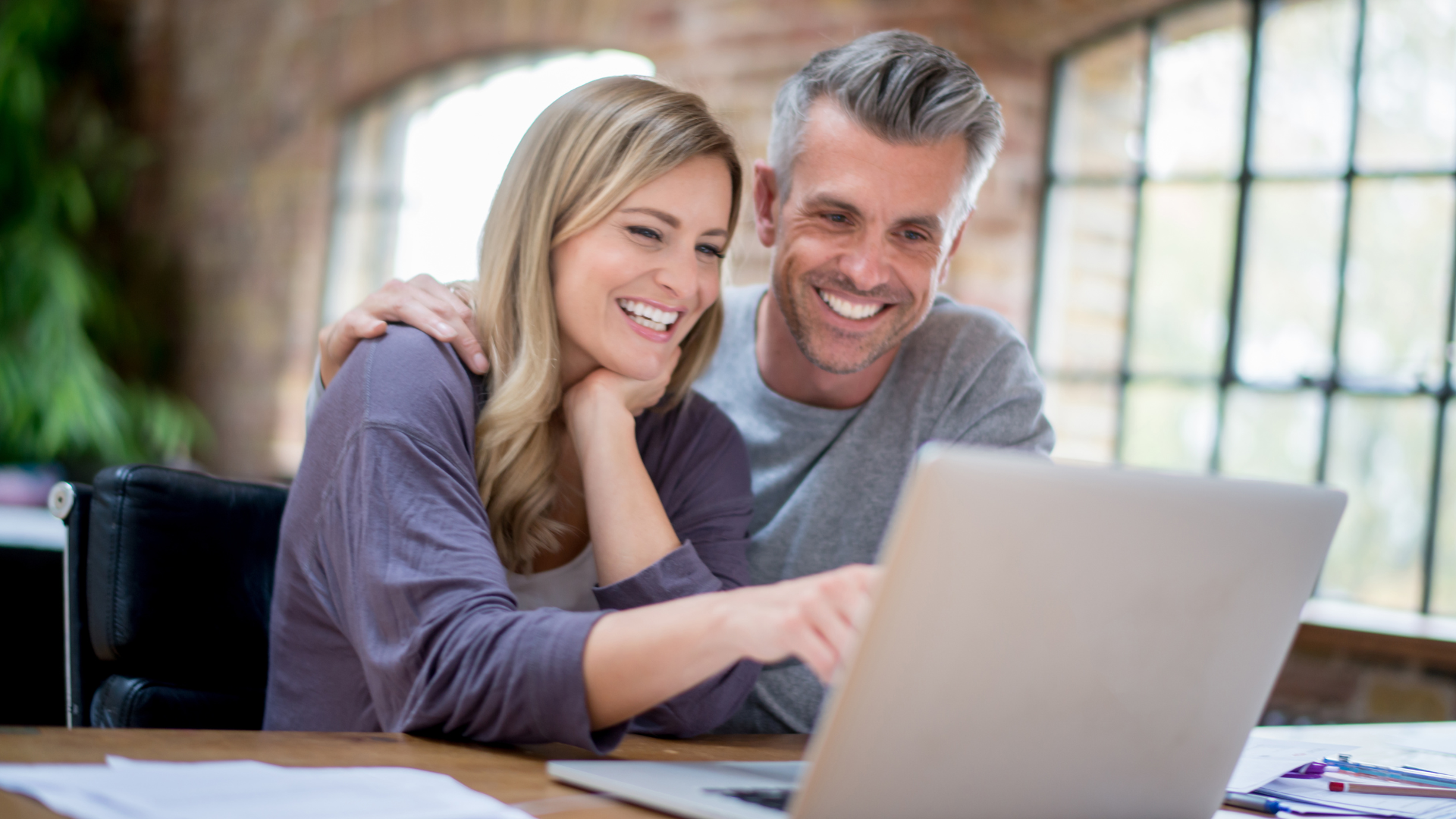 Two smiling people working together on a laptop at a table in a bright room