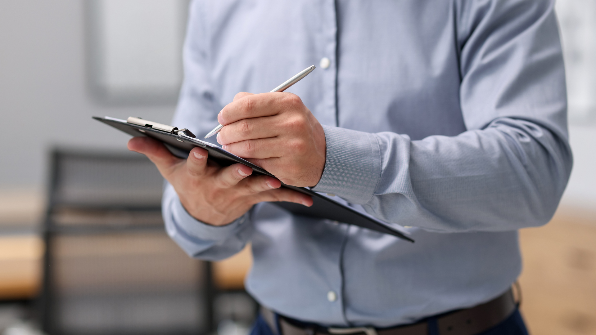 Person holding a clipboard and writing with a pen, wearing a light blue shirt indoors.