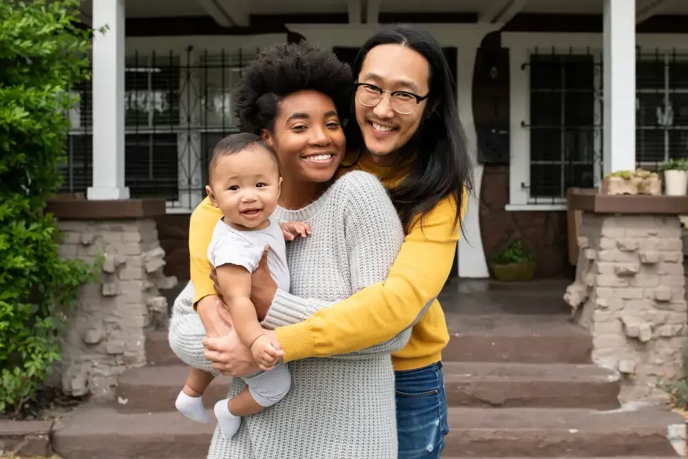 A man and a woman are holding a baby in front of a house.