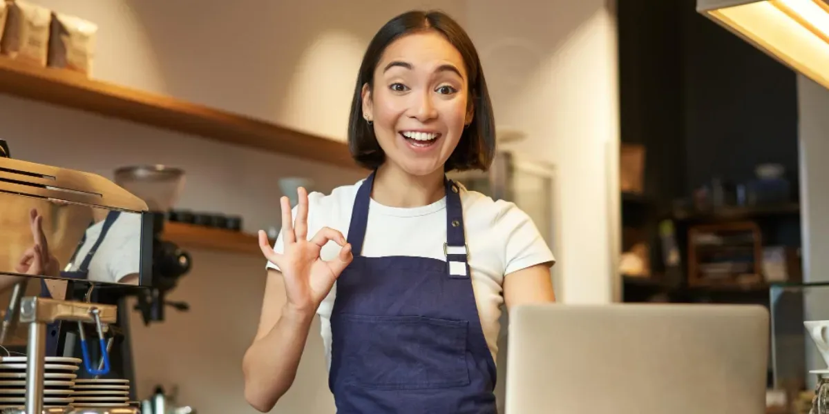A woman in an apron is giving an ok sign in a restaurant.