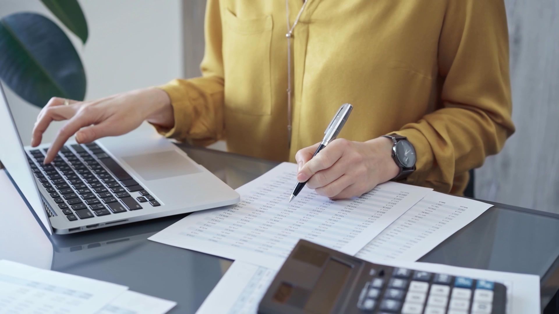 Person in yellow shirt writing on paper with laptop and calculator nearby.