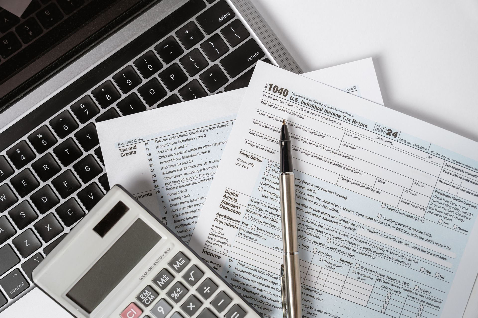 Close-up of a 1040 tax form on top of a laptop, and below a pen and calculator.