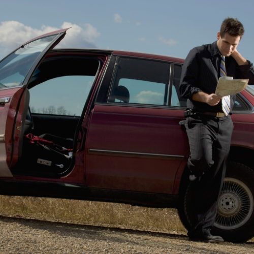 Man in suit leans against burgundy car, reading a map, door open, rural setting.