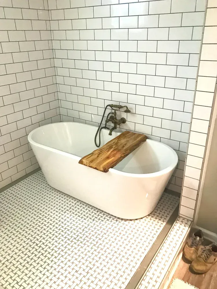 White oval bathtub in a bathroom with white brick-like tiles and a wooden tray.