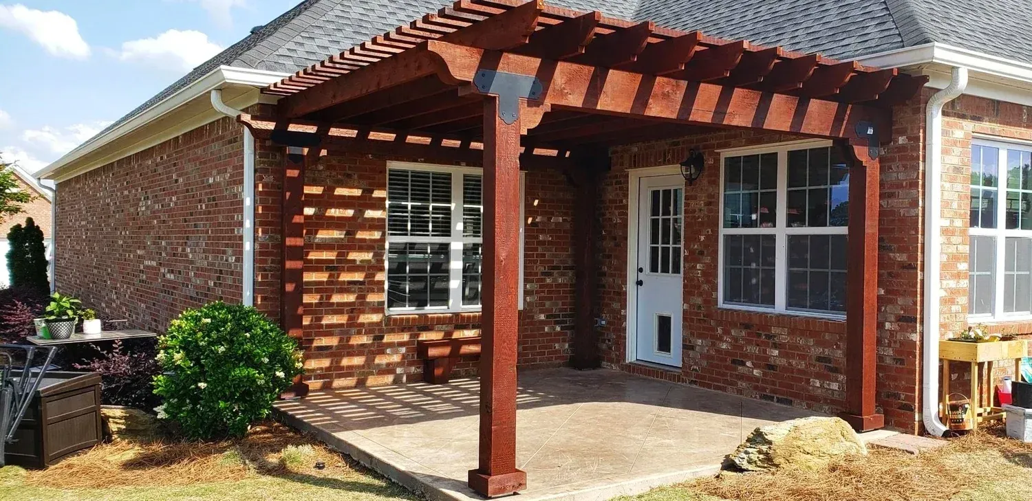 Brick house with a pergola over a patio. The pergola is dark brown. There's a door and windows.