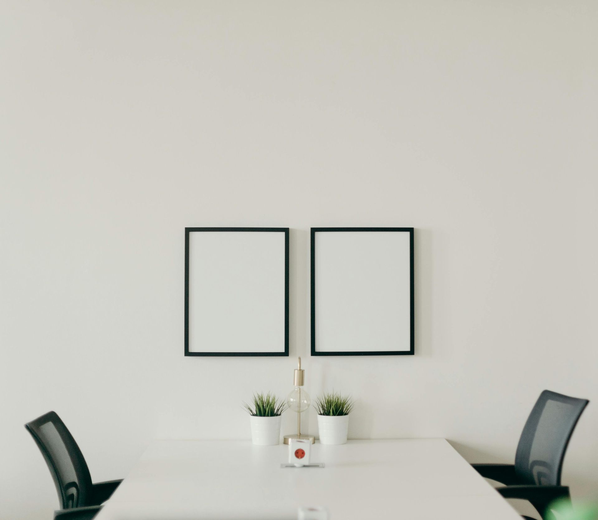 Two framed blank pictures above a table with two plants and a bottle, with two chairs.