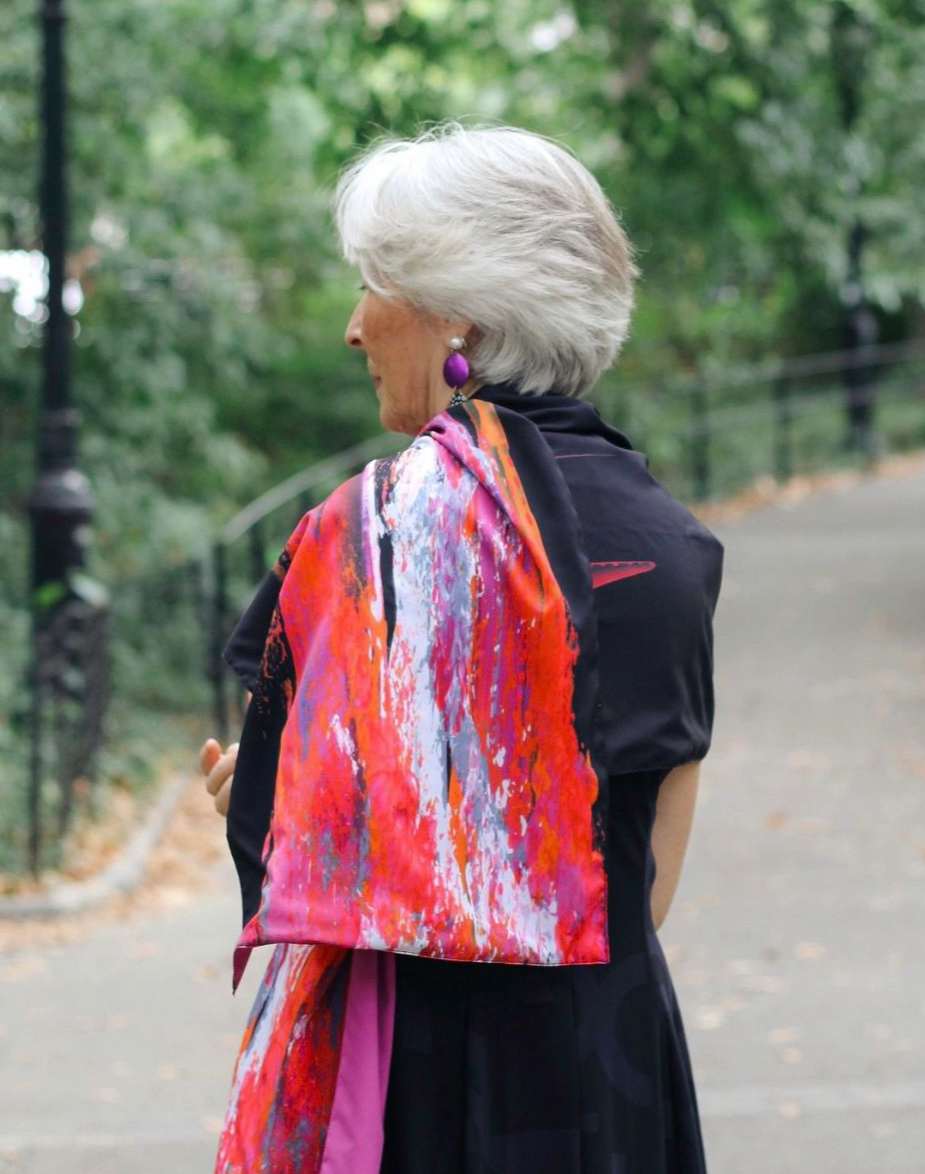 Woman with gray hair, wearing colorful scarf, purple earrings, and black dress in park.