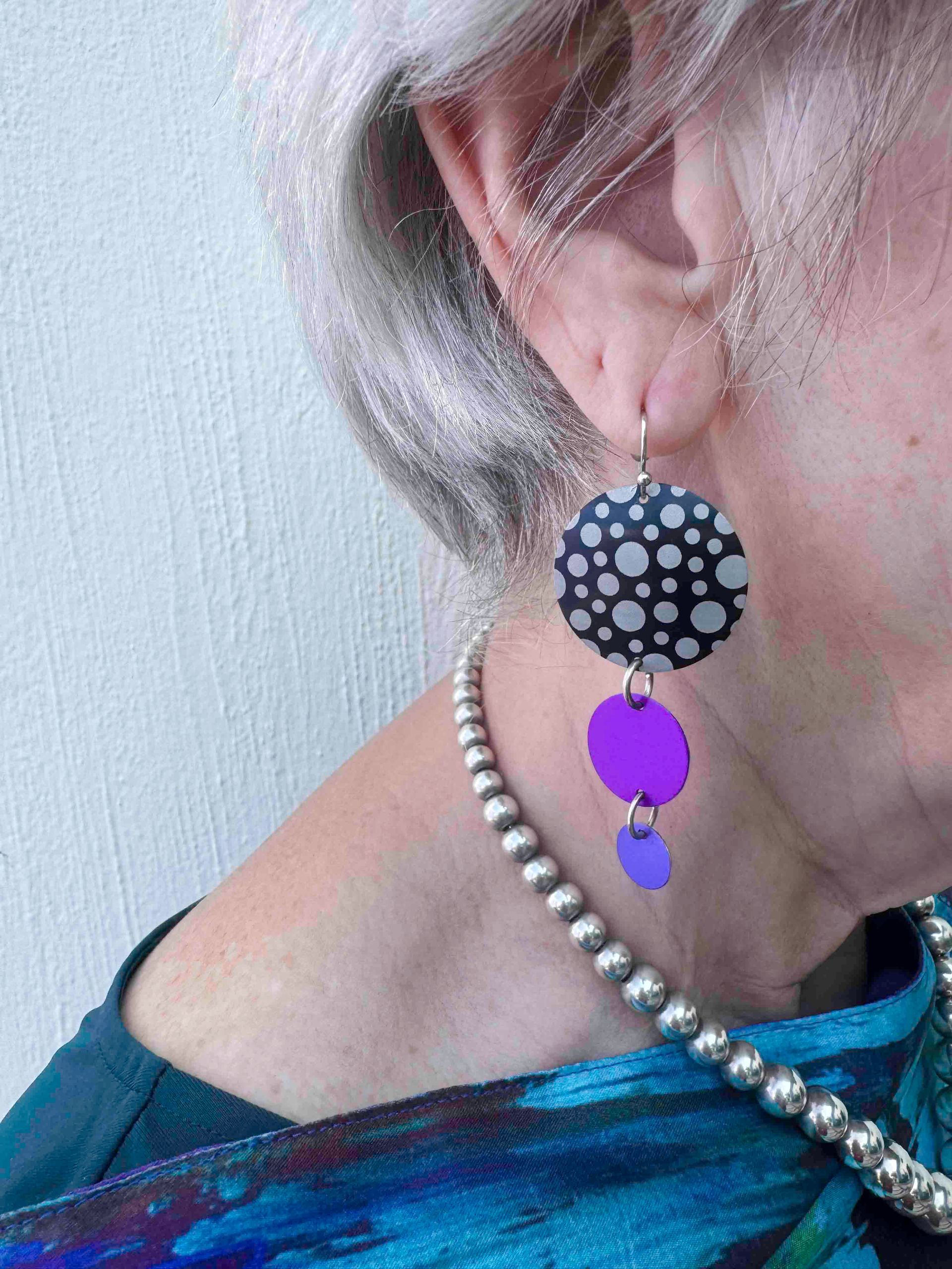 Woman wearing layered circle earrings; black and white polka dot top, purple middle, and small purple bottom with a silver necklace.