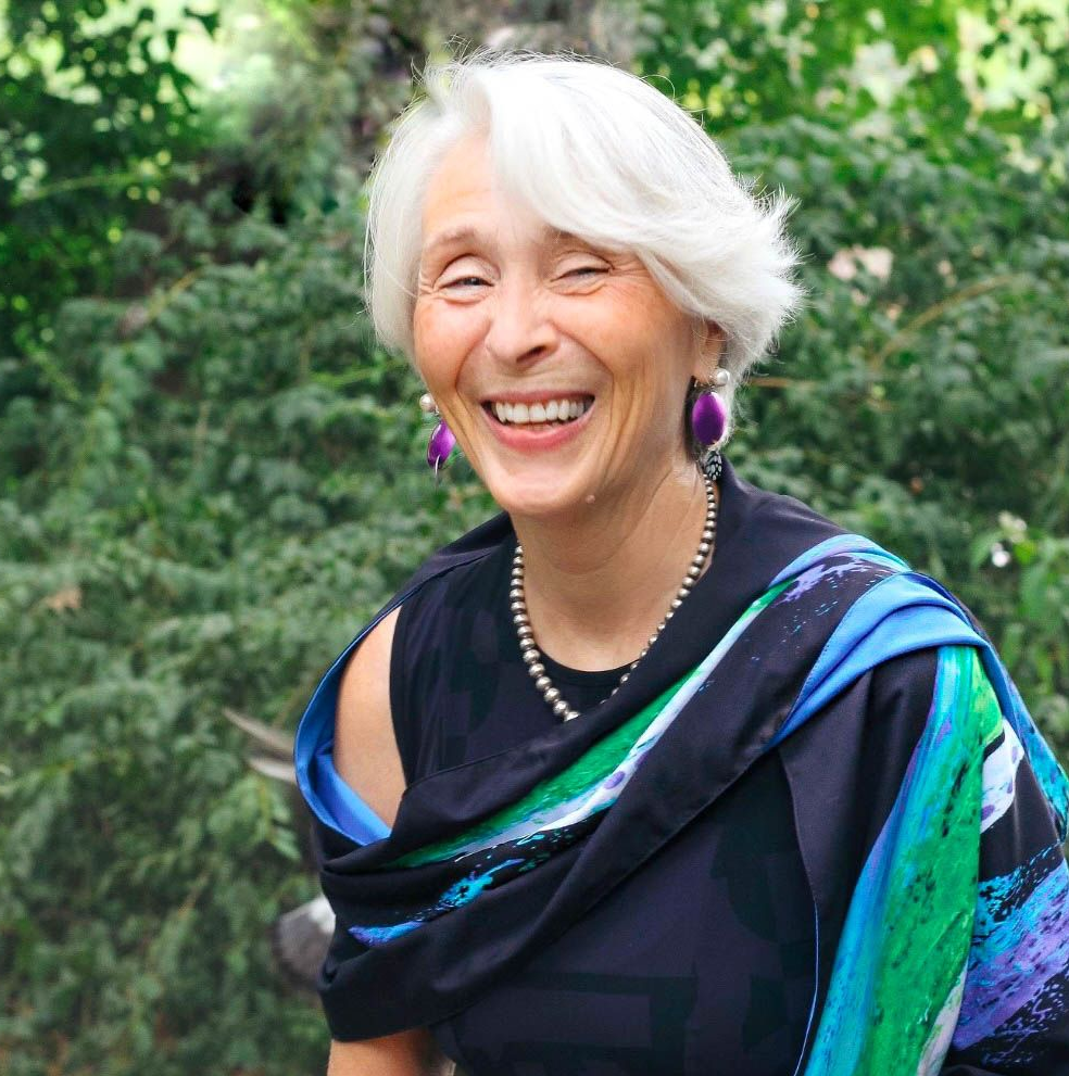 Woman with white hair smiling, wearing a black top, scarf, and purple earrings, set outdoors in front of greenery.