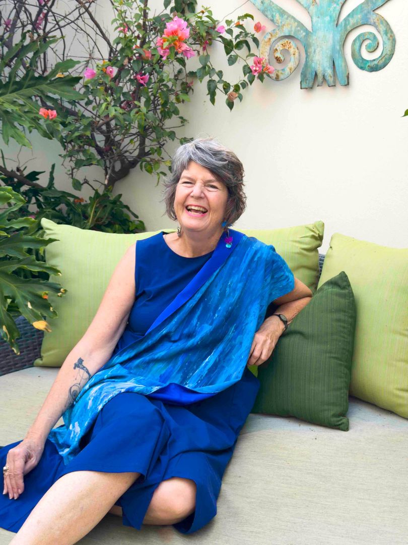 Woman in blue dress smiling on a sofa in a garden courtyard with green cushions and flowers above