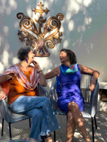 Two people sitting outdoors in colorful dresses, talking under a decorative wall ornament.