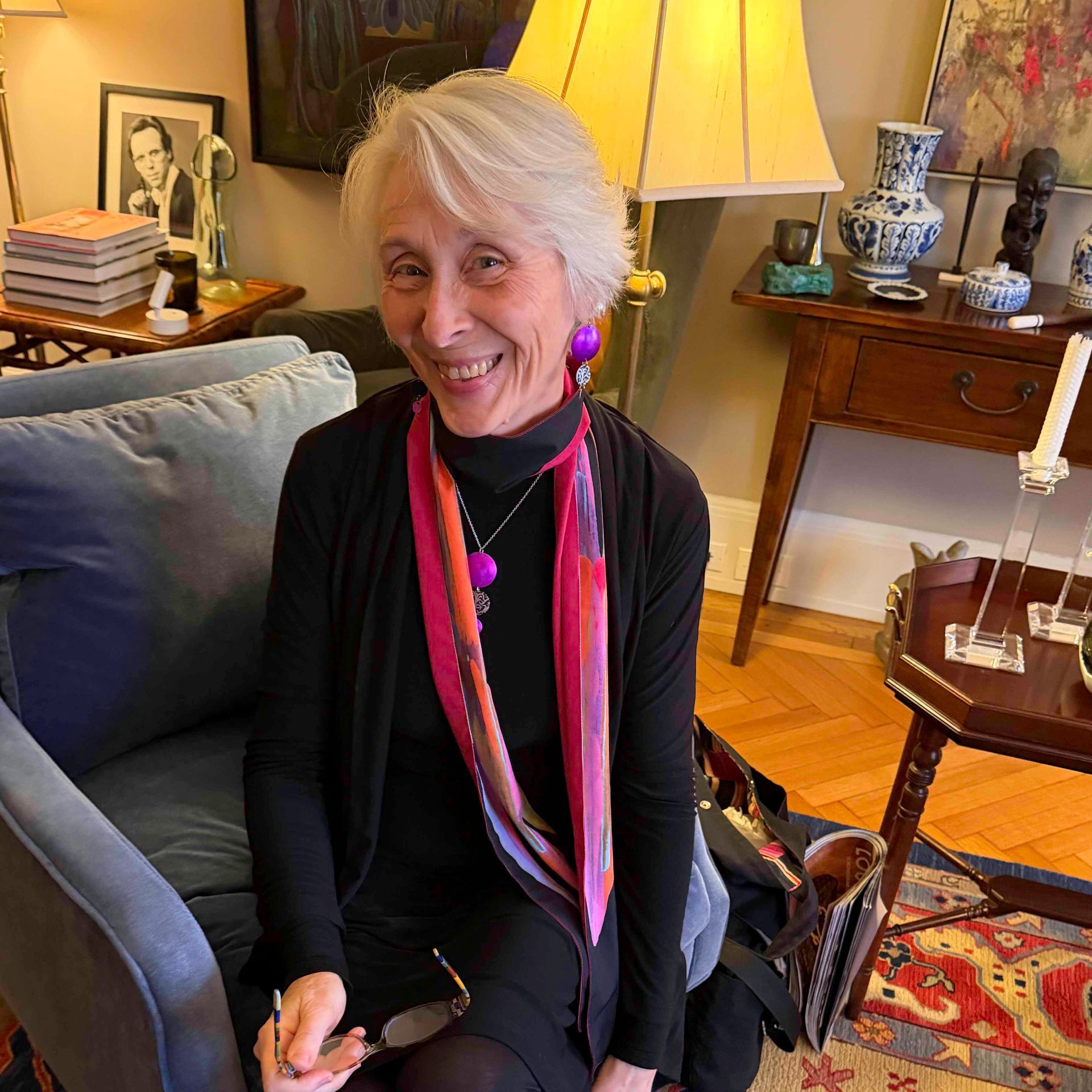 Smiling woman seated on a sofa in a living room, wearing a black outfit with a pink scarf.