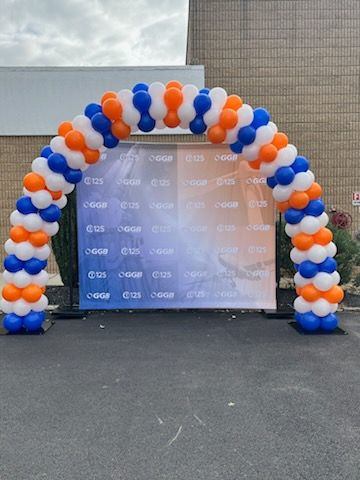 Orange, blue, and white balloon arch created by My Life In Balloons LLC for GGB’s 125th anniversary event. Arch frames a branded step-and-repeat backdrop for a photo area.