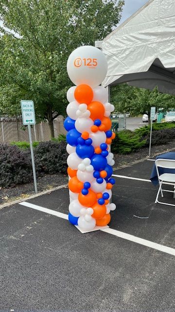 Balloon columns in orange, blue, and white created by My Life In Balloons LLC for GGB’s corporate anniversary event. Placed under a tent near dining tables for a cohesive brand look.