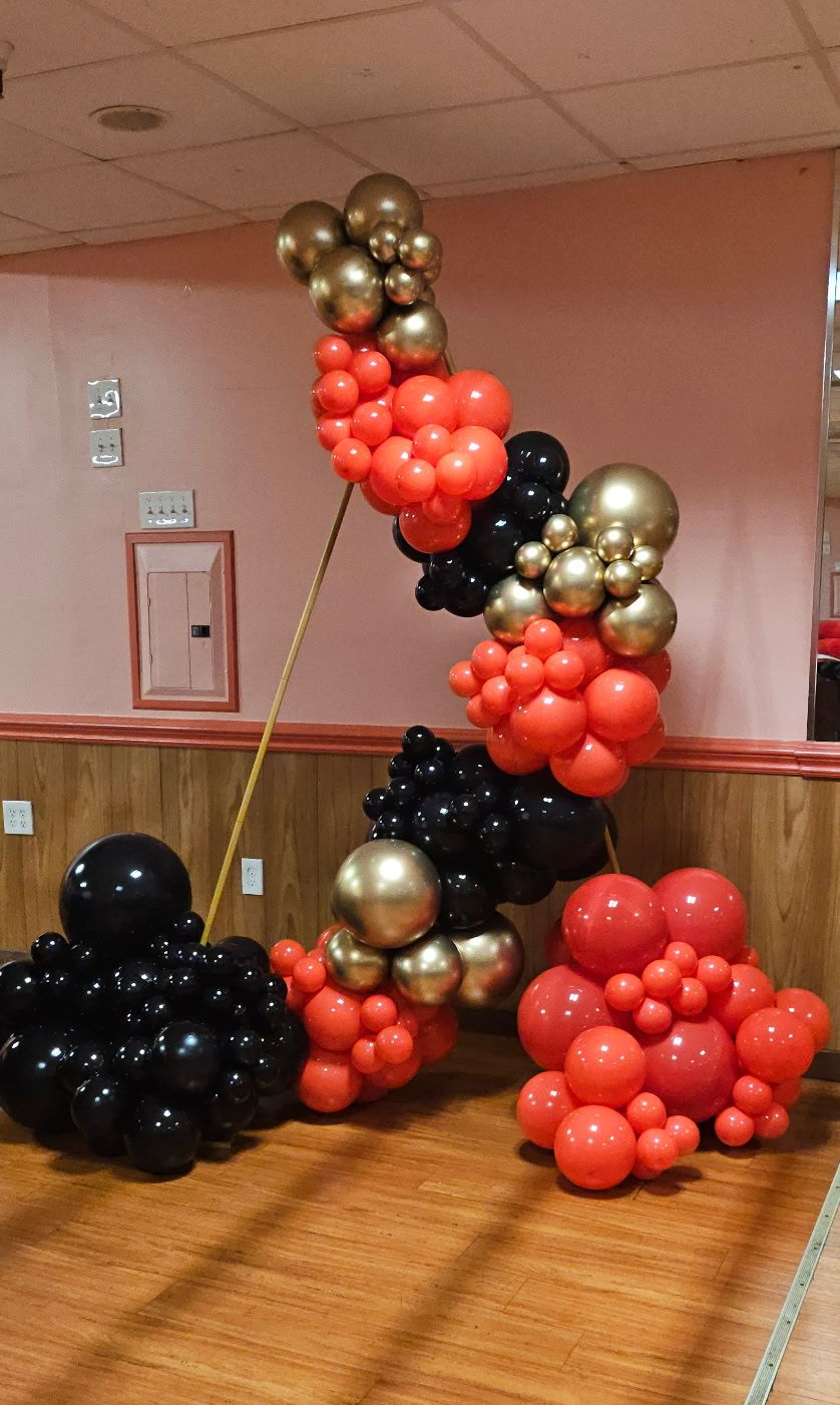 Triangle-shaped red, black, and gold balloon arch created by My Life In Balloons LLC, featuring clusters of metallic gold balloons for a striking and modern event backdrop.