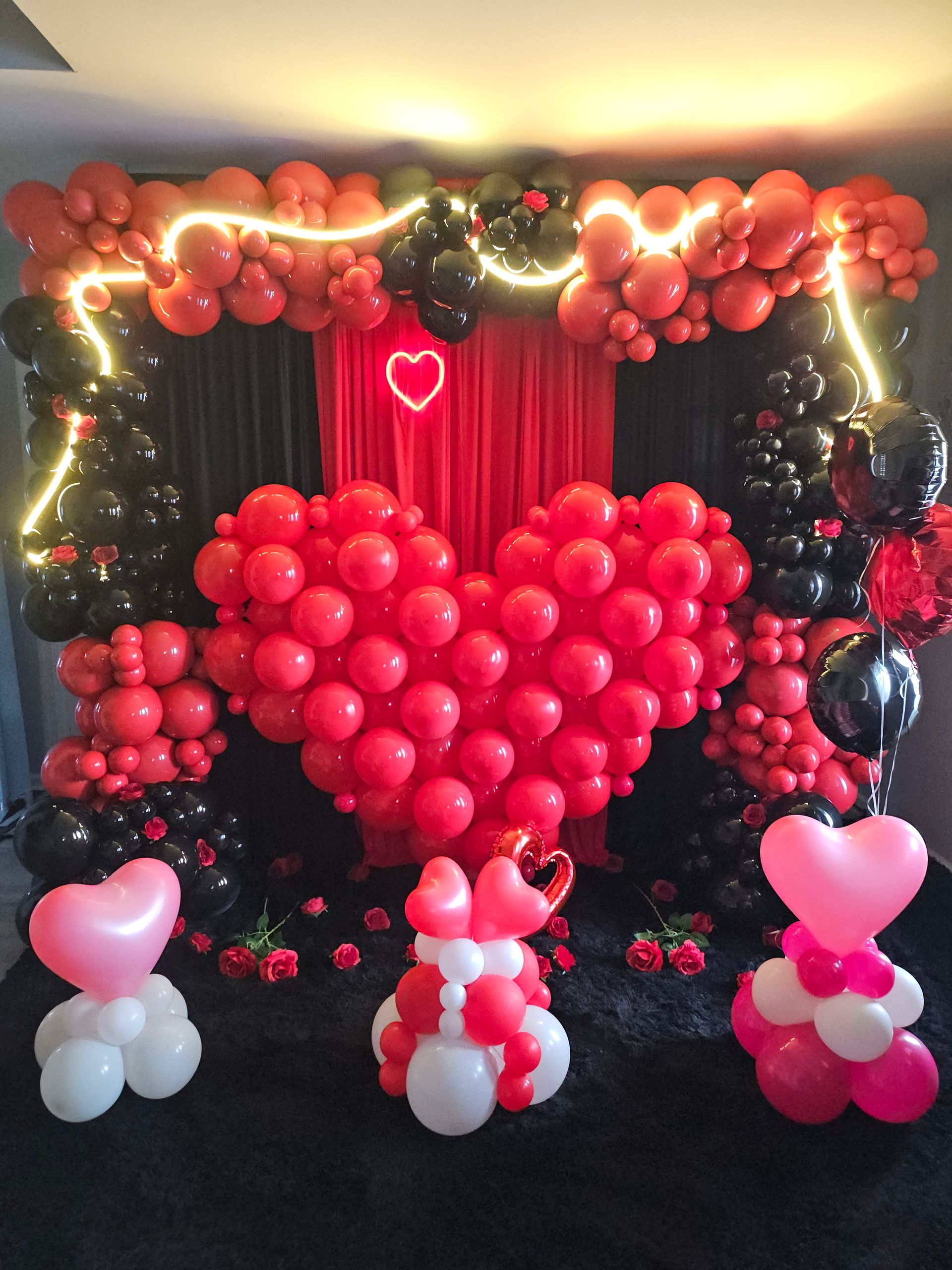 Valentine’s Day balloon decor by My Life In Balloons LLC with a large red balloon heart, black and red garlands, heart-shaped balloon pedestals, and romantic lighting.