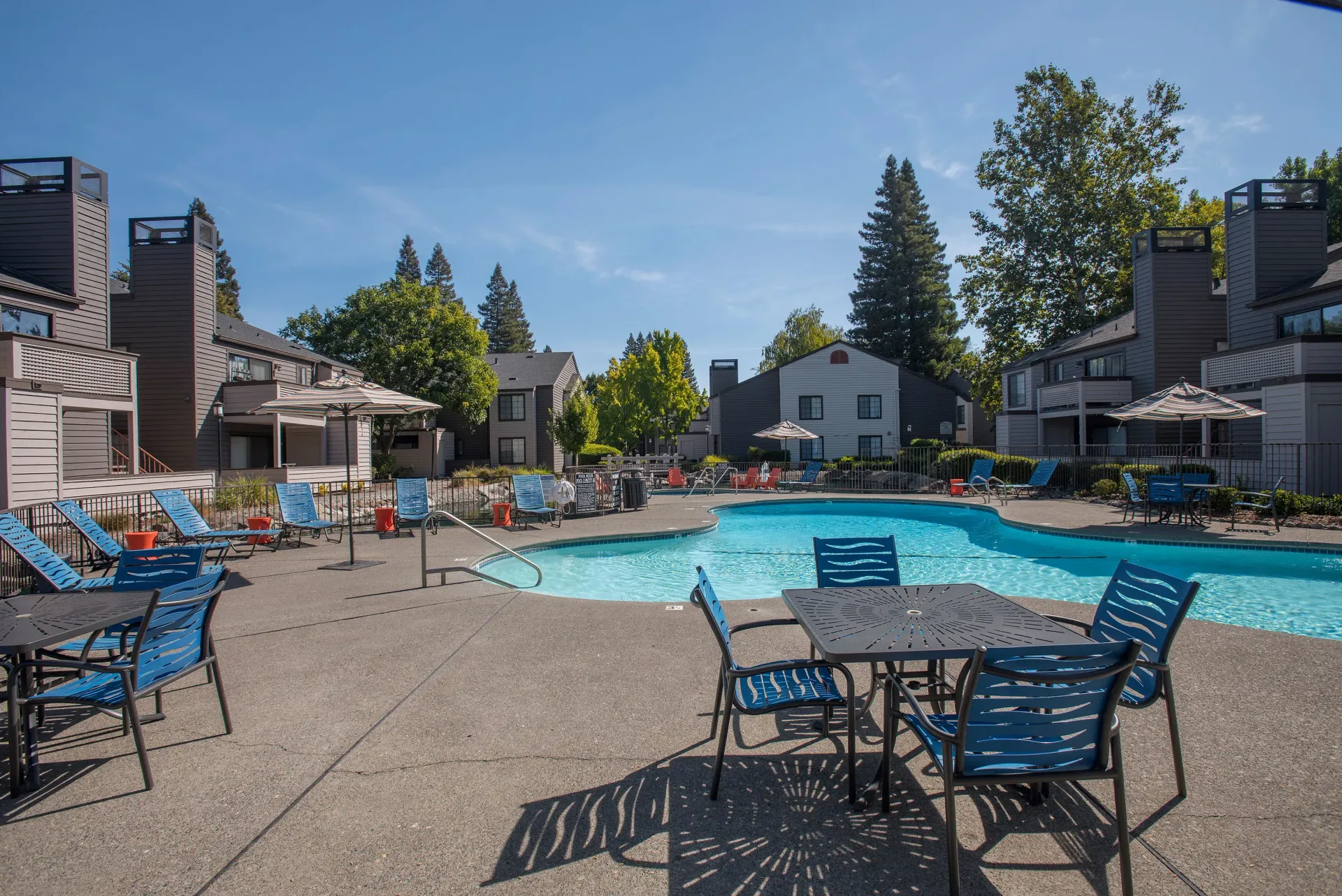 Pool area with tables, chairs, and lounge chairs. Apartment buildings and trees in the background. Blue sky at Hidden Lake apartments in Sacramento, CA.