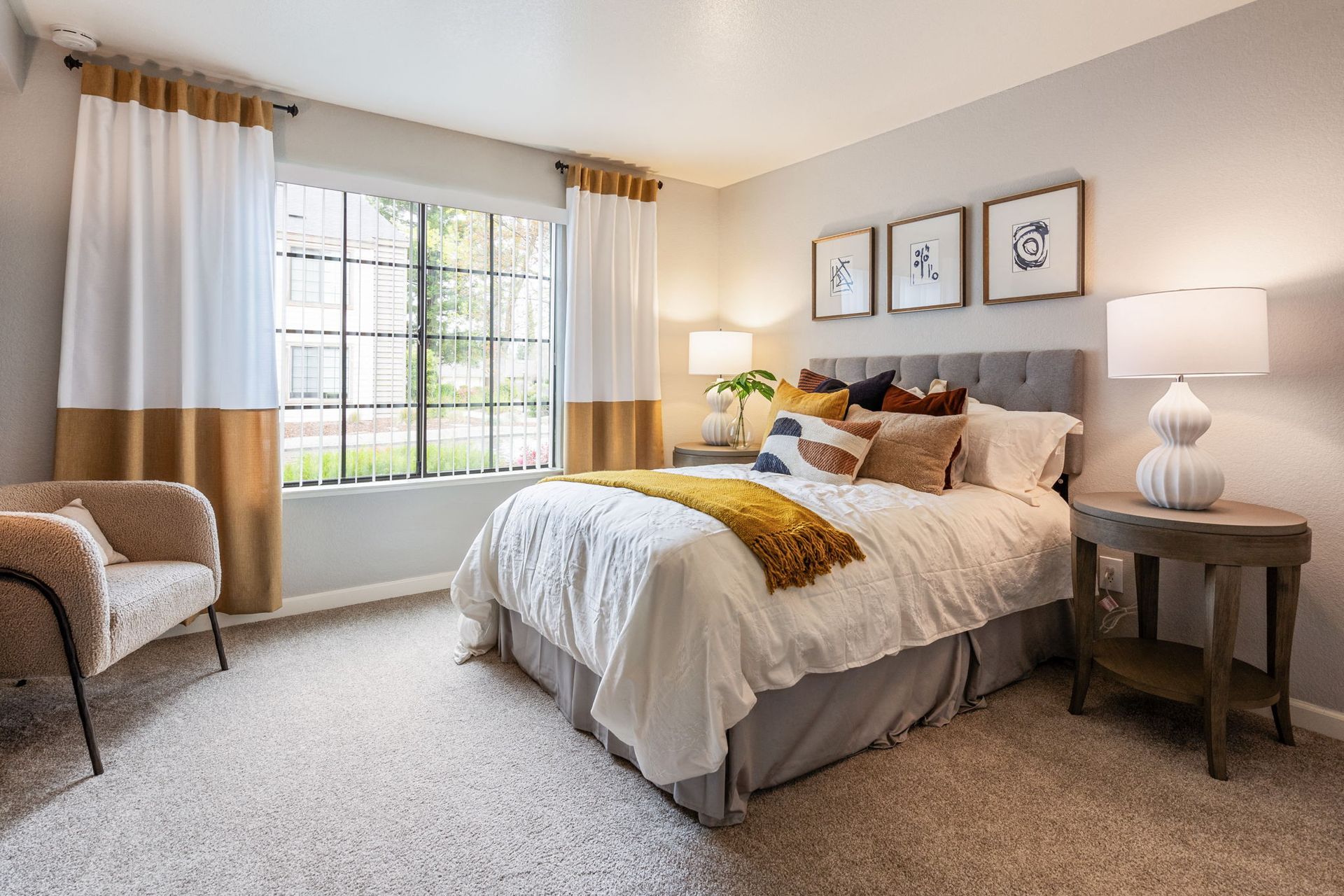 Bedroom with a plush bed, two nightstands, lamps, and a large window with two-tone curtains at Hidden Lake apartments in Sacramento, CA.