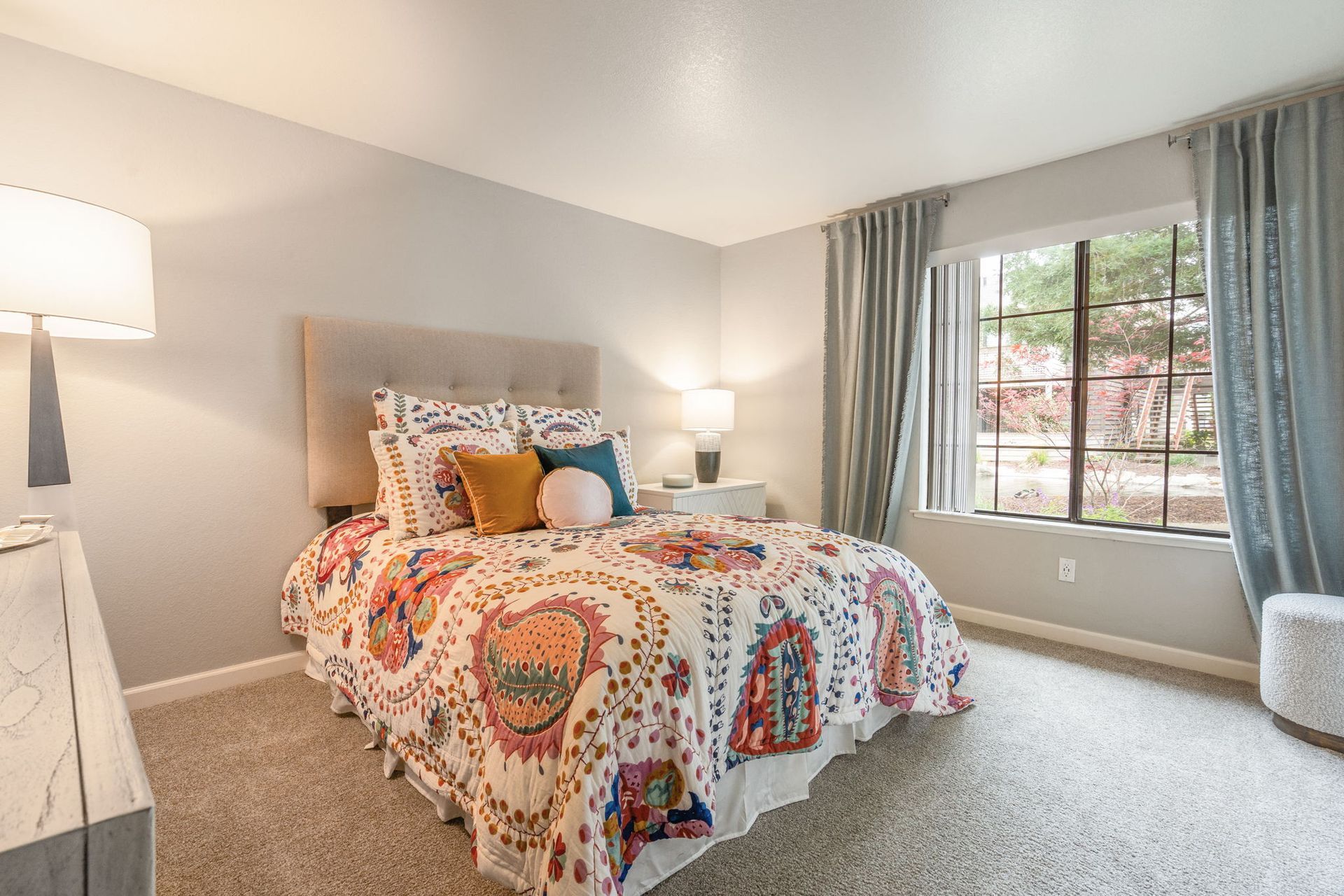 Bedroom in apartment with queen bed, colorful quilt, two nightstands, and large window with blue curtains at Hidden Lake apartments in Sacramento, CA.