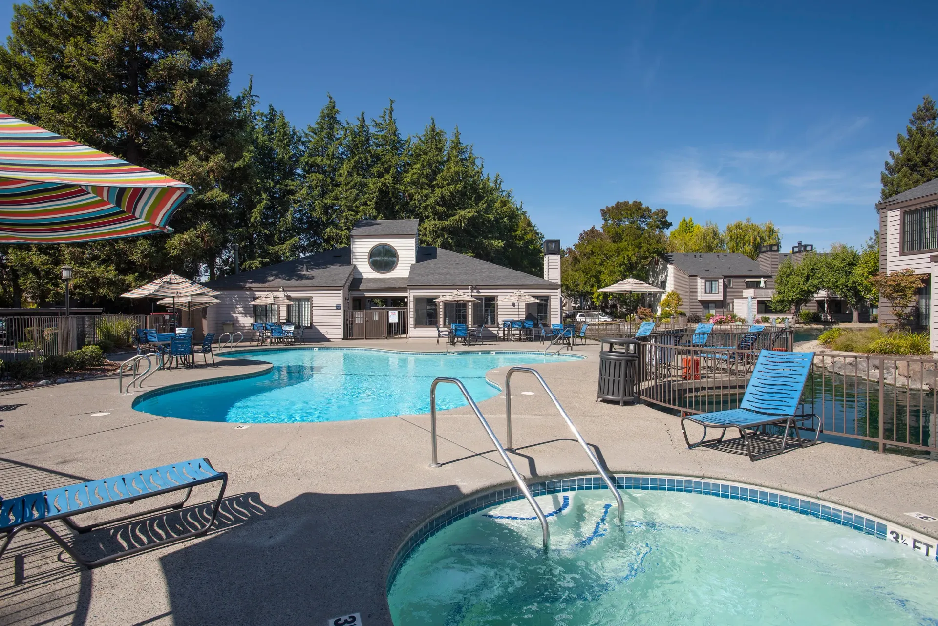 Pool and jacuzzi at an apartment complex with blue lounge chairs and umbrellas at Hidden Lake apartments in Sacramento, CA.