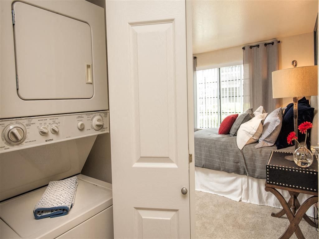 Stacked washer and dryer in a closet beside a bedroom with a bed and nightstand at Hidden Lake apartments in Sacramento, CA.